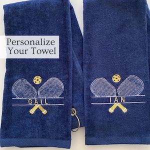 Peut inclure: Deux serviettes personnalisées bleu marine avec des motifs brodés de raquettes et de balles de tennis. Les serviettes ont des accents dorés et les noms "GAIL" et "IAN" brodés. Chaque serviette a une boucle de suspension dorée.