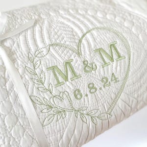 May include: White quilted blanket featuring a heart-shaped design embroidered in green. The embroidery includes the initials "M & M" and the date "6.8.24". A white ribbon is tied around the blanket. The blanket has a textured, quilted pattern.