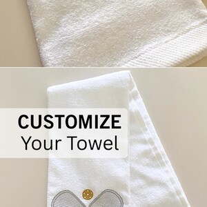 May include: White custom pickleball towel with embroidered pickleball paddles, ball, and the name "TRICIA" in pink. The towel has a loop for hanging and the text "CUSTOM PICKLEBALL TOWEL" is at the top.