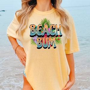 Puede incluir: Una camiseta amarilla con un estampado gr&aacute;fico de las palabras "Beach Bum" en un estilo retro con hojas y flores tropicales.