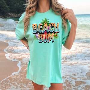 Puede incluir: Una camiseta azul turquesa con el texto "Beach Bum" en una fuente retro con un dise&ntilde;o de flores tropicales.