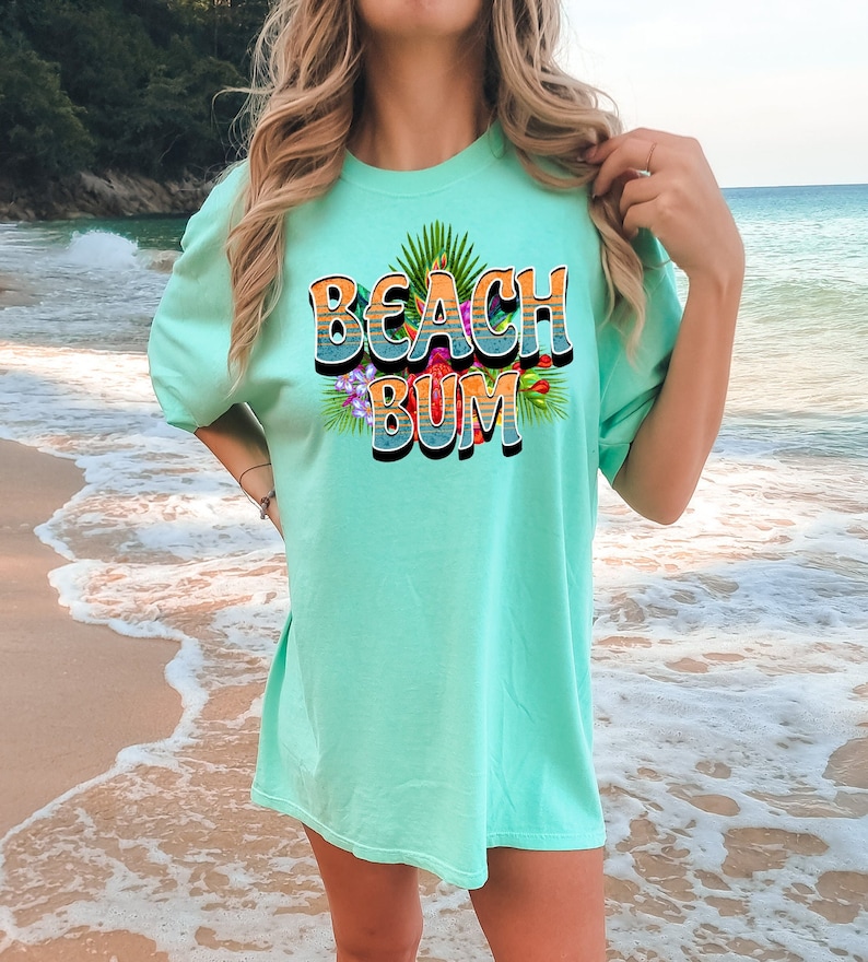 Puede incluir: Una camiseta azul turquesa con el texto "Beach Bum" en una fuente retro con un dise&ntilde;o de flores tropicales.