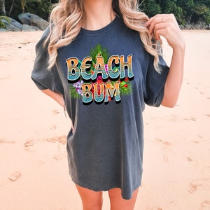 Puede incluir: Una camiseta gris oscuro con un gr&aacute;fico colorido que dice "Beach Bum" con flores y hojas tropicales.