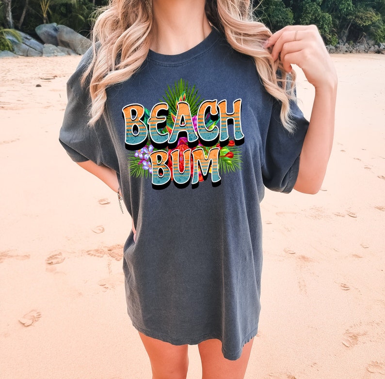 Puede incluir: Una camiseta gris oscuro con un gr&aacute;fico colorido que dice "Beach Bum" con flores y hojas tropicales.