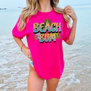 Puede incluir: Una camiseta rosa con el texto "Beach Bum" en un estilo retro con flores coloridas y hojas de palma alrededor del texto.