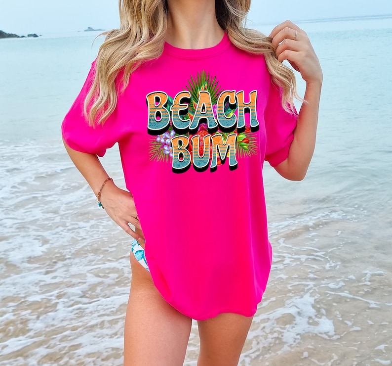 Puede incluir: Una camiseta rosa con el texto "Beach Bum" en un estilo retro con flores coloridas y hojas de palma alrededor del texto.