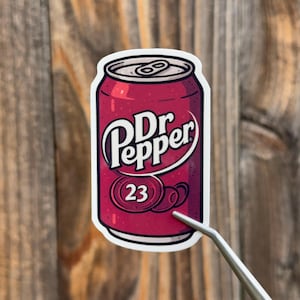 Op de afbeelding: Een sticker met een gestileerde afbeelding van een Dr Pepper frisdrankblikje. Het blikje is dieprood met het Dr Pepper-logo in wit en het nummer 23. De sticker wordt vastgehouden door een zilveren pincet.