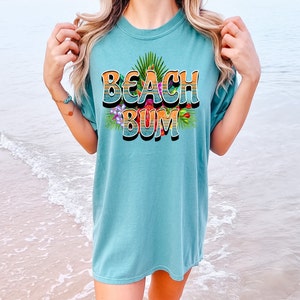Puede incluir: Una camiseta azul turquesa con un estampado gr&aacute;fico de las palabras "Beach Bum" en un estilo retro con rayas naranja y amarillas y flores y hojas tropicales.
