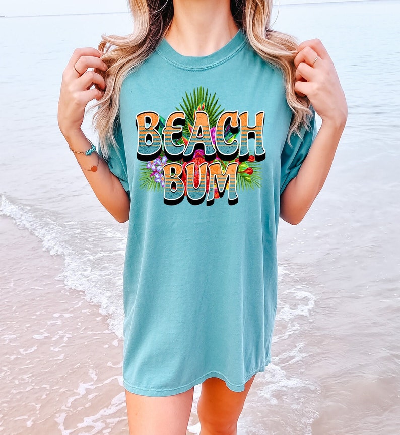 Puede incluir: Una camiseta azul turquesa con un estampado gr&aacute;fico de las palabras "Beach Bum" en un estilo retro con rayas naranja y amarillas y flores y hojas tropicales.