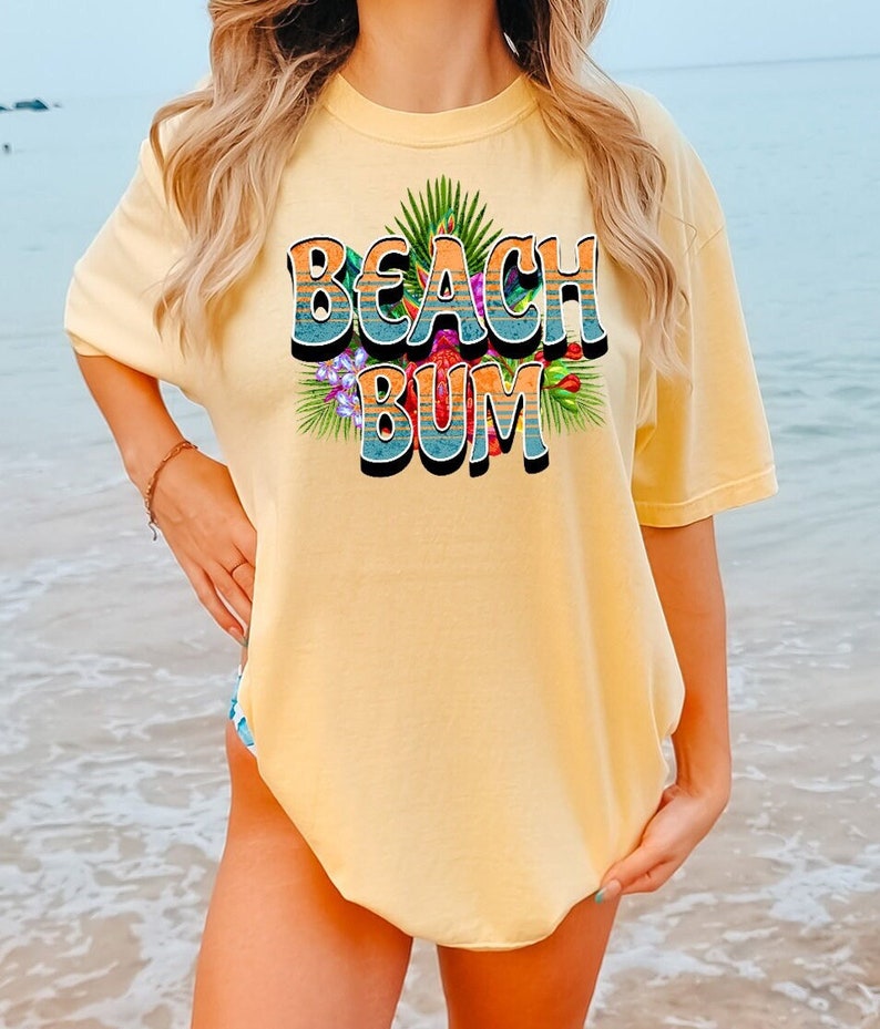 Puede incluir: Una camiseta amarilla con un estampado gr&aacute;fico de las palabras "Beach Bum" en un estilo retro con hojas y flores tropicales.