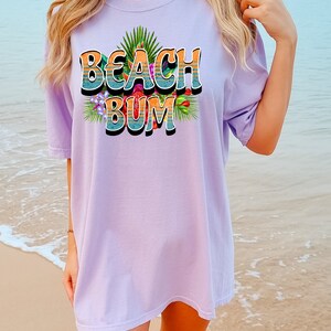 Puede incluir: Una camiseta de gran tama&ntilde;o de color lavanda con un estampado gr&aacute;fico del texto "Beach Bum" en un estilo retro con flores coloridas y hojas de palmera.