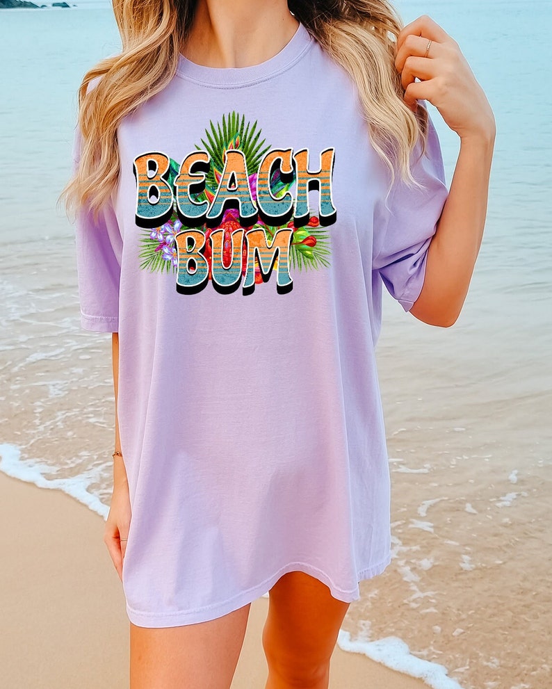 Puede incluir: Una camiseta de gran tama&ntilde;o de color lavanda con un estampado gr&aacute;fico del texto "Beach Bum" en un estilo retro con flores coloridas y hojas de palmera.
