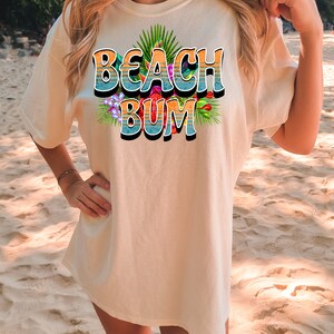 Puede incluir: Una camiseta blanca con un estampado gr&aacute;fico del texto "Beach Bum" en letras azules y naranjas con flores y hojas tropicales.