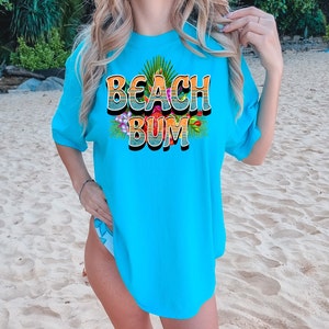 Puede incluir: Una camiseta azul turquesa con un estampado gr&aacute;fico de las palabras "Beach Bum" en un estilo retro con flores coloridas y hojas de palma.