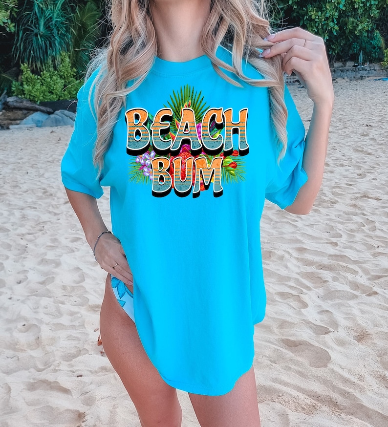 Puede incluir: Una camiseta azul turquesa con un estampado gr&aacute;fico de las palabras "Beach Bum" en un estilo retro con flores coloridas y hojas de palma.