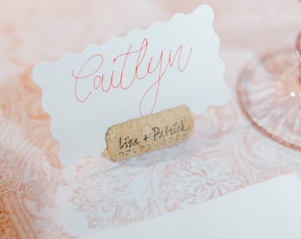 May include: White place card with the name "Caitlyn" written in red cursive. The place card is held up by a cork with the text "Liza + Patrick 02.01.2020" written on it.