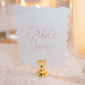 Könnte beinhalten: Eine weiße Karte mit gewellten Rändern und einem goldenen Metallständer. Auf der Karte steht in rosa Kursivschrift "Table Seven".