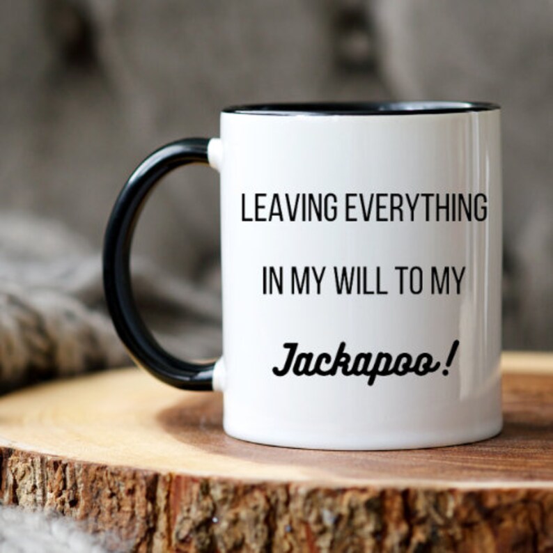 Pu&ograve; includere: Tazza in ceramica bianca con manico e bordo neri. La tazza presenta il testo "LEAVING EVERYTHING IN MY WILL TO MY Jackapoo!" in lettere nere. La tazza &egrave; appoggiata su una superficie di legno.