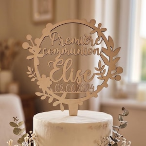 Può includere: Una torta bianca a due piani con decorazioni floreali e un cake topper in legno. Il topper recita "Première communion Elise 02.06.2024". La torta è su un supporto di legno, circondata da candele e verde.