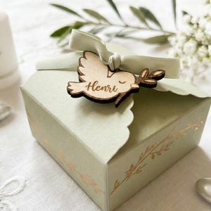 Può includere: Una scatola regalo verde chiaro con bordo smerlato, ornata da un ornamento a colomba in legno e un nastro verde chiaro. La colomba ha inciso il nome "Henri". La scatola è decorata con accenti floreali dorati.