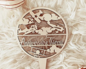 May include: A wooden cake topper with a round design featuring airplanes and clouds. The text "Anniversaire Thierry" is written on the topper.