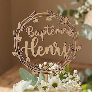 Peut inclure: Un gâteau à deux étages, recouvert de crème au beurre blanche, orné d'une décoration en bois avec l'inscription "Baptême Henri". Décoré de fleurs blanches et de verdure, posé sur un présentoir en bois. Bougies et fleurs en arrière-plan.