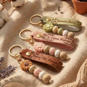 Peut inclure: Trois porte-clés en cuir avec des détails dorés et des perles. Chaque porte-clés a une lanière en cuir de couleur différente avec des motifs floraux et du texte français : "Maman Chérie", "Je t'aime Maman" et "Bonne Fête Maman".