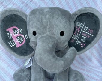 May include: Gray plush elephant with black ears. The ears have a chalkboard surface with pink lettering. The left ear says "Brooke Elizabeth". The right ear says "Tuesday August 24, 2020 7:02 oz 21.5 in".