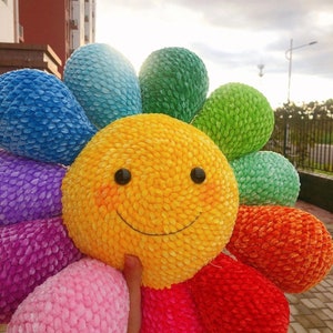 Puede incluir: Una flor de peluche colorida con una cara amarilla sonriente. La flor tiene pétalos en tonos de azul, verde, morado, rosa, rojo y naranja.