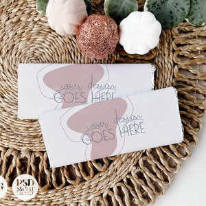 May include: Two white chocolate bar wrappers with a pink abstract design and the text "your design goes here". The wrappers are on a woven brown mat with decorative plants and white pumpkins.