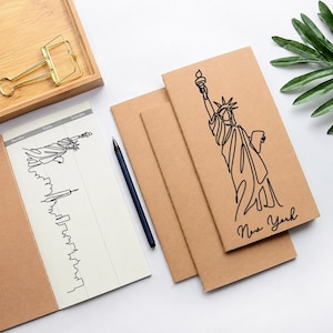 Könnte beinhalten: Drei hellbraune Papiernotizbücher mit einer schwarzen Strichzeichnung der Freiheitsstatue und dem Text New York auf dem Einband. Ein Notizbuch ist geöffnet und zeigt eine schwarze Strichzeichnung der Skyline von New York und der Freiheitsstatue.