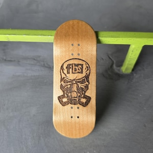 May include: A light brown wooden fingerboard deck featuring a dark brown laser-engraved graphic of a skull wearing a gas mask with the text fbs centered on the forehead. The deck includes four sets of pre-drilled truck mounting holes.