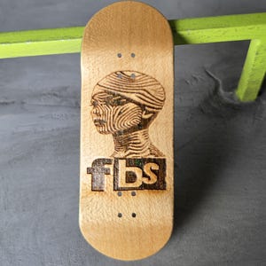 May include: A light-colored wooden fingerboard deck featuring a dark, laser-engraved graphic of a human head in profile with intricate line patterns. The text fbs is engraved in a bold, blocky font below the head. The deck has four sets of holes.