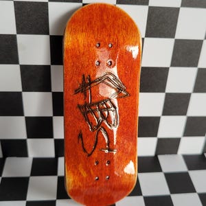 May include: A fingerboard with a red-brown stain finish and a black graphic of a bird with an arrow on the bottom. The fingerboard is on a black and white checkered background.