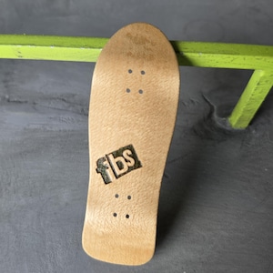 May include: A light brown wooden fingerboard deck featuring a natural wood grain texture. The deck has four sets of pre-drilled truck mounting holes and a black square logo with the letters fbs printed in white on the center of the board surface.