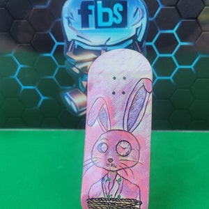 May include: A pink fingerboard with a graphic of a pink rabbit wearing a suit and tied up with rope. The rabbit has one eye covered with a bandage and the other eye is open. The fingerboard has three holes for the trucks.