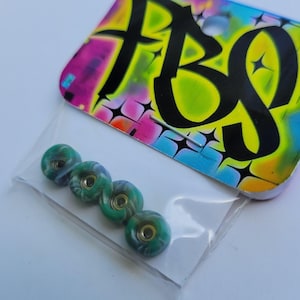 May include: Four small, green and blue swirl skateboard wheels in a clear plastic bag. The wheels have a gold-colored center. A colorful tag with the letters "FBG" is visible in the background.