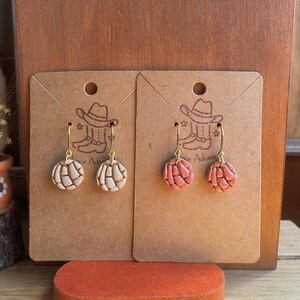 May include: Two pairs of handmade earrings displayed on brown cards. One pair is light beige, the other is pink. Each earring has a gold-colored hook and a round, textured design. The cards have a cowboy boot and hat illustration and the word "Adorn".