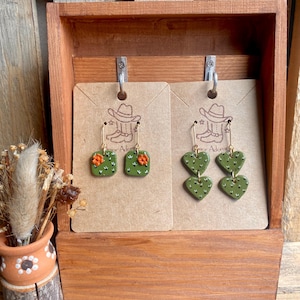 May include: Two pairs of handmade earrings displayed in a wooden box. One pair features square green cactus designs with orange flowers, and the other has heart-shaped green cactus designs. Both pairs have gold-colored ear wires.