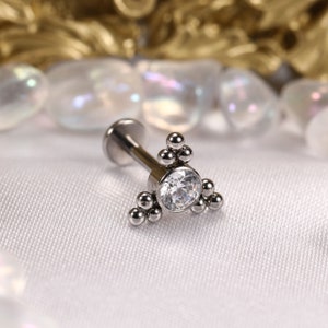 May include: A silver body piercing with a clear crystal and a cluster of small silver beads in a triangular shape.