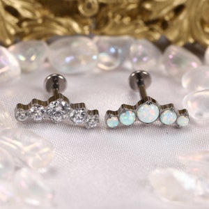 May include: Two pieces of jewelry, each with a silver-colored base and a row of gemstones. One features clear, round stones, while the other has opalescent, oval-shaped stones. The jewelry is set against a white background.