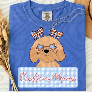 Może przedstawiać: Niebieska koszulka Comfort Colors z wizerunkiem kreskówkowego psa w okularach przeciwsłonecznych w kształcie gwiazd i patriotycznych kokardkach. Koszulka ma etykietę w kratkę z napisem "Custom Name" kursywą. Srebrny naszyjnik jest zarzucony na koszulkę.