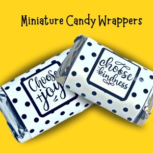 May include: Two miniature candy wrappers with a white background and navy blue polka dots. One wrapper says "Choose Joy" and the other says "Choose Kindness" in a handwritten font. The wrappers are on a yellow background.
