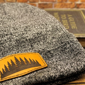 Pode incluir: Um gorro de malha cinza e branco com um patch de couro com uma silhueta de floresta preta. O patch é retangular com cantos arredondados e é costurado no chapéu. O chapéu é dobrado na aba.