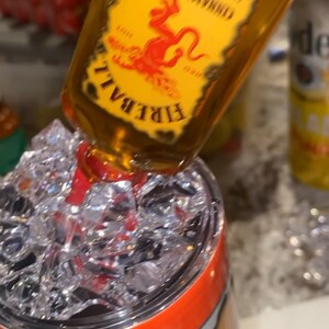 Op de afbeelding: Een fles Fireball Cinnamon Whisky die in een roestvrijstalen beker met ijs giet. De beker heeft een oranje en geel ontwerp met het woord "Fireball" erop.