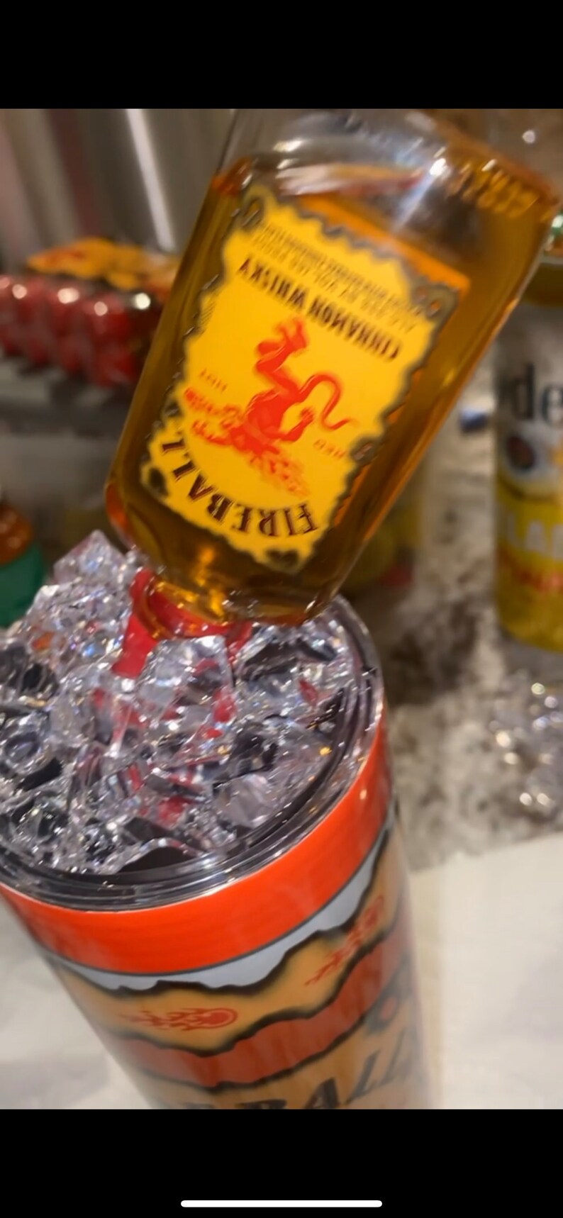 Op de afbeelding: Een fles Fireball Cinnamon Whisky die in een roestvrijstalen beker met ijs giet. De beker heeft een oranje en geel ontwerp met het woord "Fireball" erop.