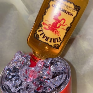 Op de afbeelding: Een glazen fles Fireball Cinnamon Whisky wordt in een tumbler met ijs gegoten. De fles heeft een rood en geel etiket met een rode duivelafbeelding. De tumbler heeft een rood en oranje ontwerp.