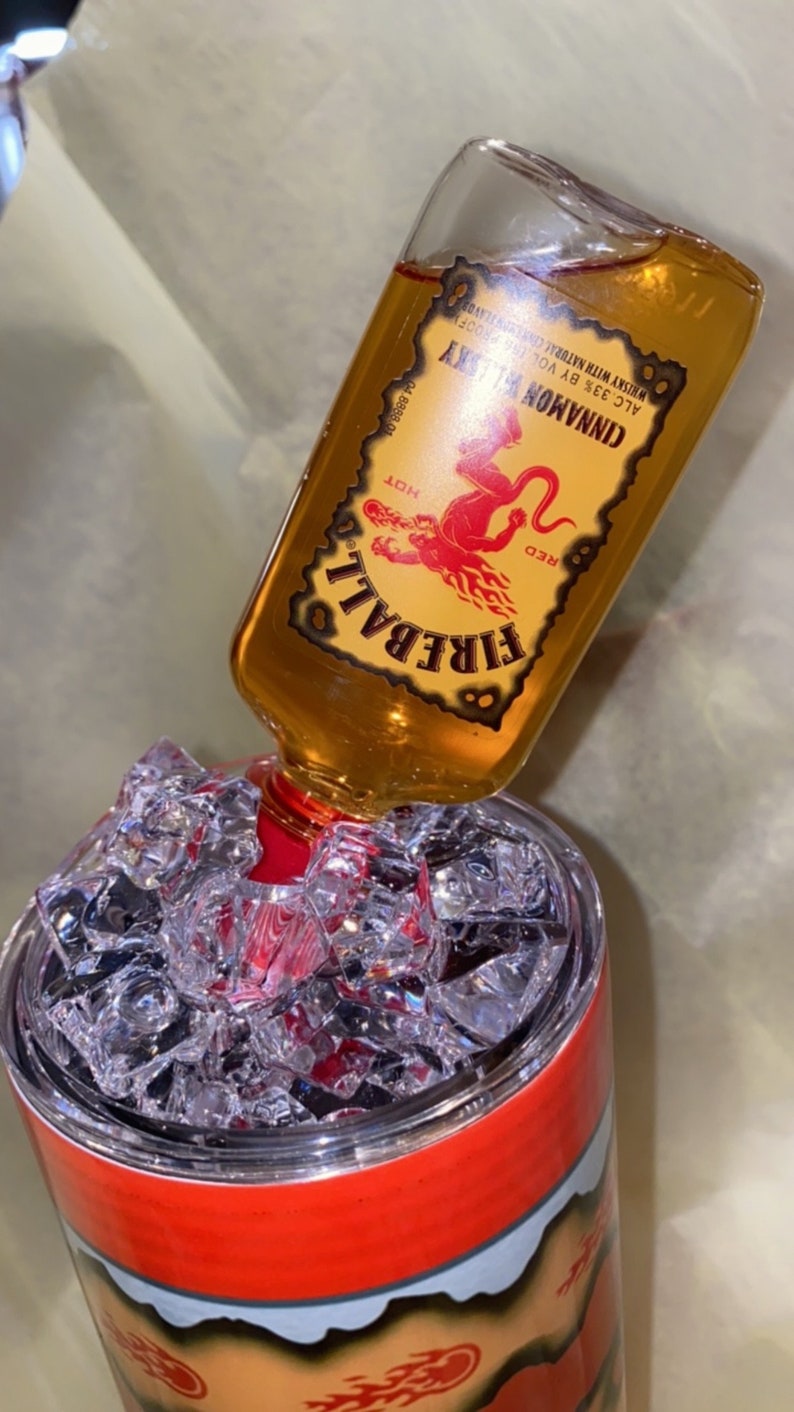 Op de afbeelding: Een glazen fles Fireball Cinnamon Whisky wordt in een tumbler met ijs gegoten. De fles heeft een rood en geel etiket met een rode duivelafbeelding. De tumbler heeft een rood en oranje ontwerp.