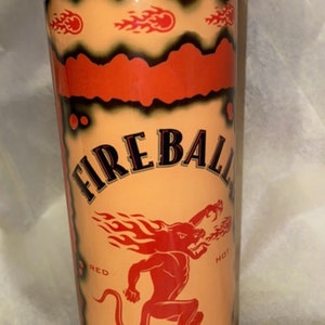 Op de afbeelding: Een cilindrische beker met een rode deksel en een ontwerp met de tekst "FIREBALL" en een rode duivelsfiguur. De beker heeft een lichtoranje achtergrond met vlammen en de woorden "CINNAMON WHISKY".