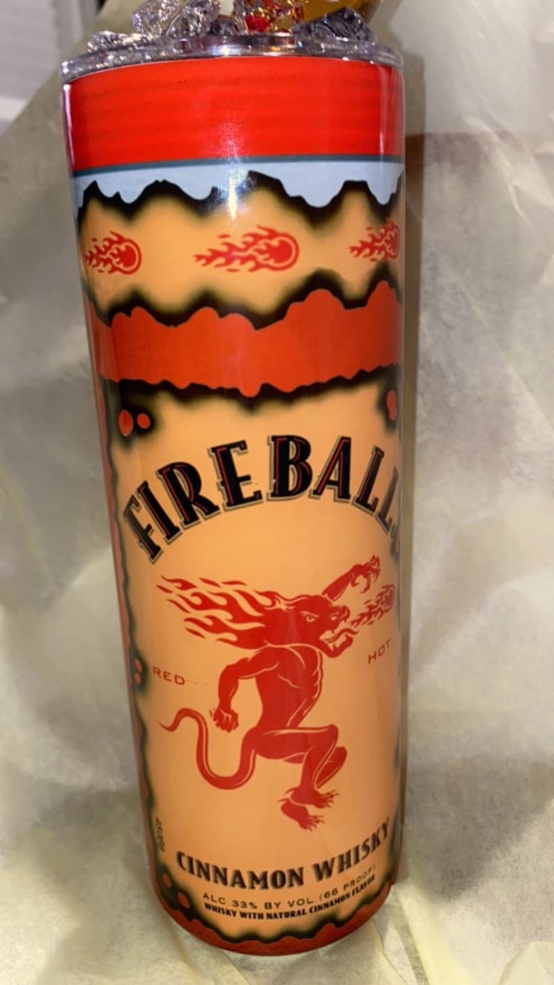 Op de afbeelding: Een cilindrische beker met een rode deksel en een ontwerp met de tekst "FIREBALL" en een rode duivelsfiguur. De beker heeft een lichtoranje achtergrond met vlammen en de woorden "CINNAMON WHISKY".